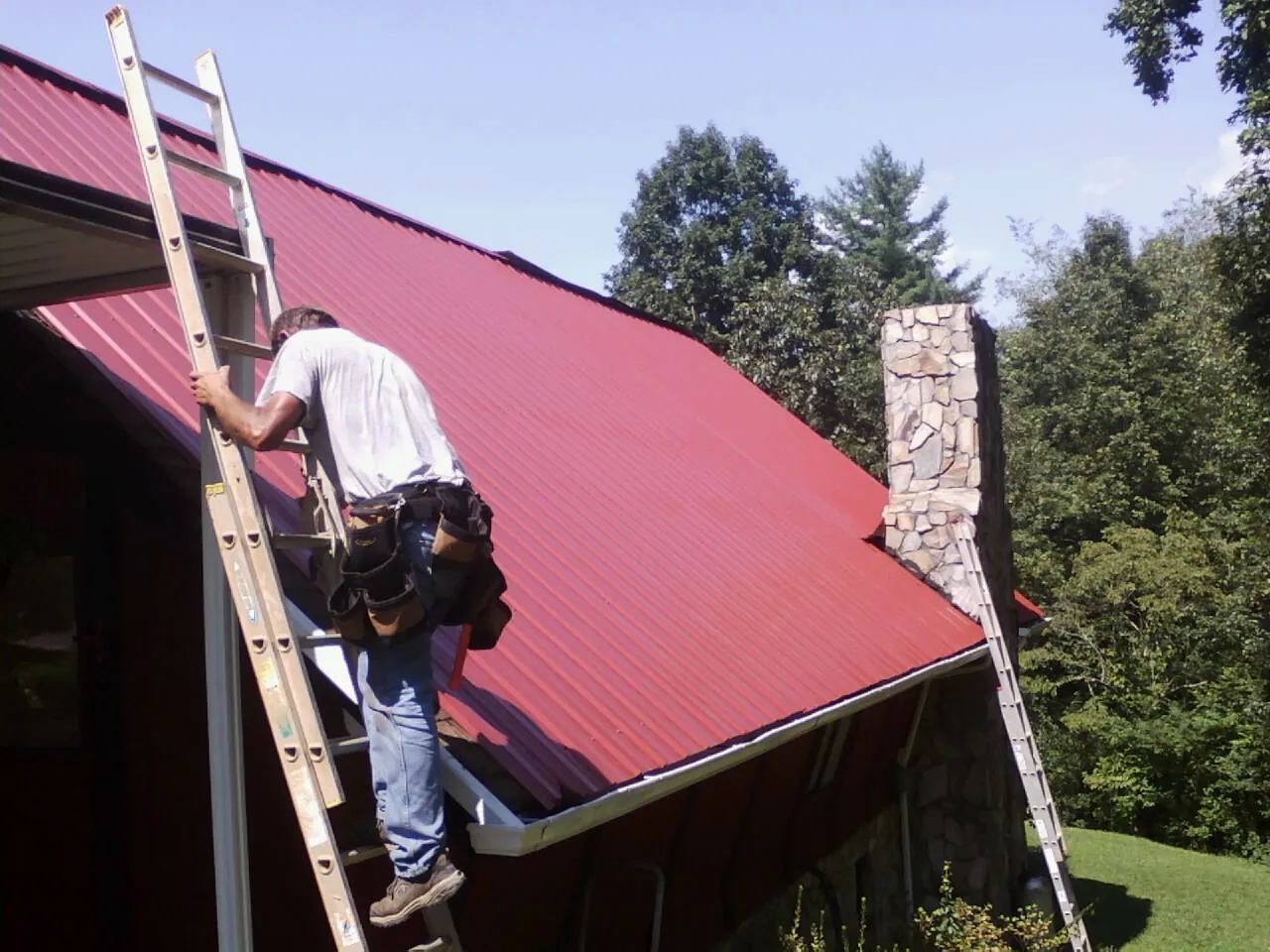 Roofer on a ladder beside a red metal roof and stone chimney — Soffit & Fascia Repair by Roofing Brothers in Blue Point